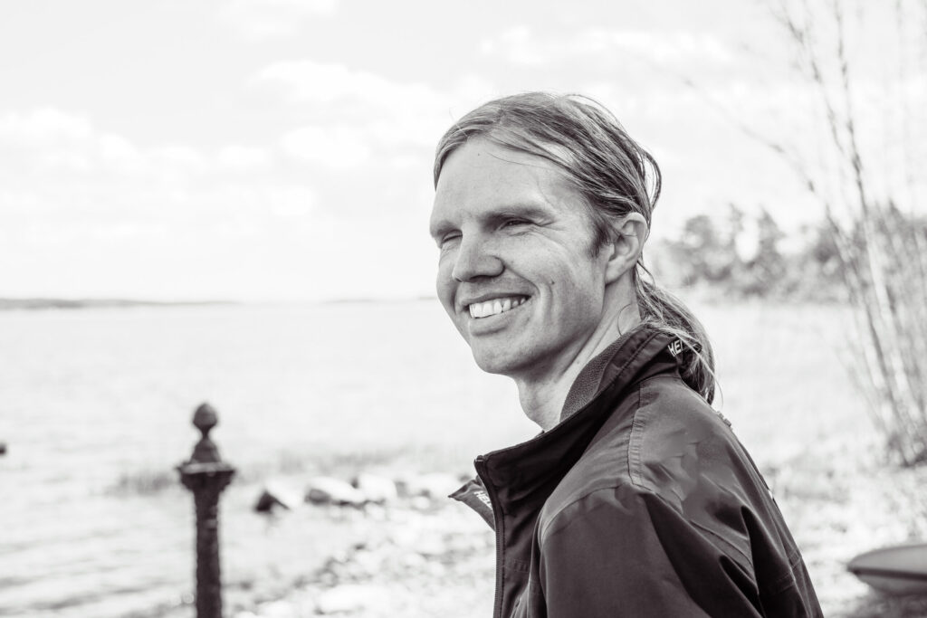 Lars has long light hair in a ponytail. He stands askew to the camera and smiles broadly wearing a dark windbreaker. The photo was taken at a lake in summertime